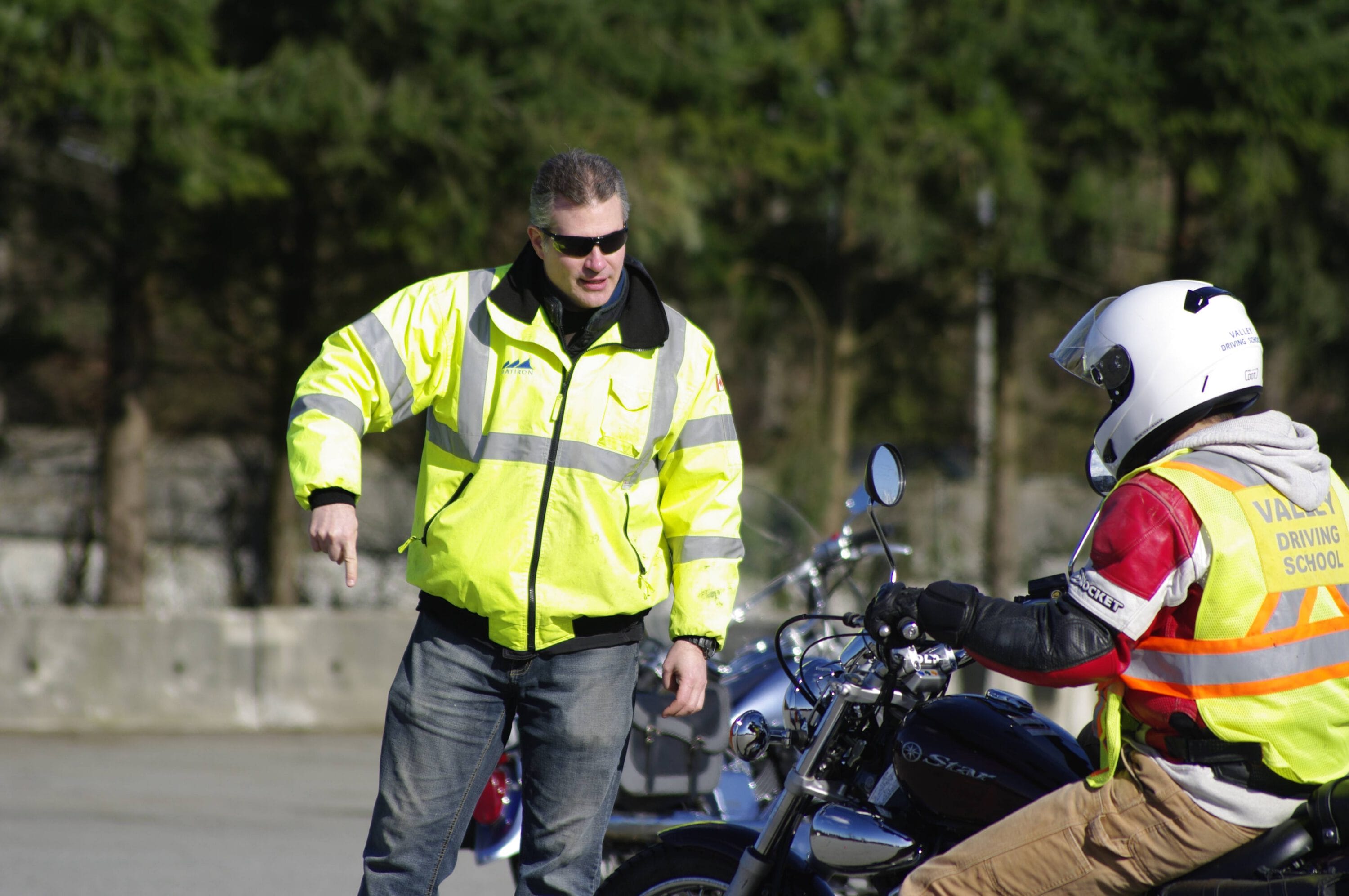 A man wearing a yellow jacket is instructing a motorcycle rider in a parking lot.