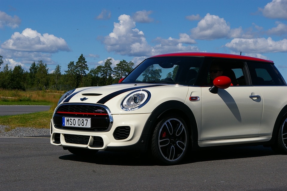 A side-view of a white and black Mini Cooper with a red roof and red mirrors, parked on an asphalt surface with a line of trees and a blue sky in the background.
