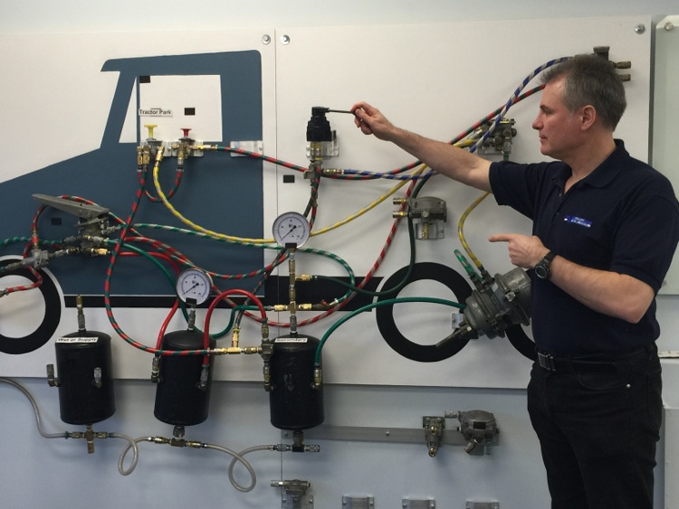 A driving instructor pointing to a semi-truck air brake system training board featuring color-coded air lines, pressure gauges, and air tanks.