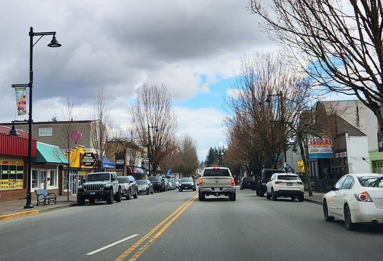 Downtown Aldergrove road with a pick up truck and car driving on the road. There is local businesses and parked cars lining the roadway.