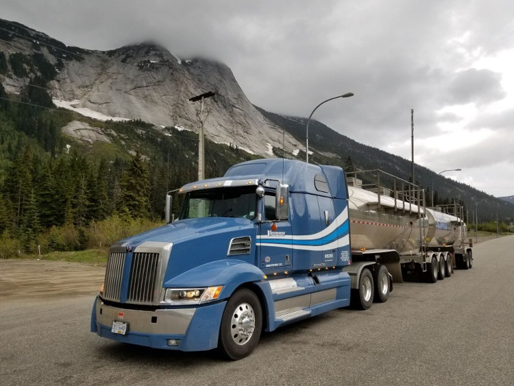 A blue Western Star semi-truck pulling double tanker trailers parked on a mountain pass in British Columbia, with steep rocky peaks and evergreen trees under a cloudy sky.