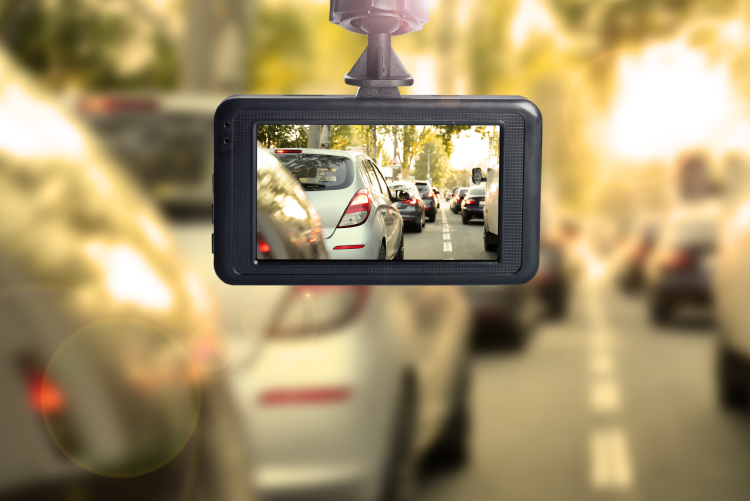 A dashcam mounted on a vehicle's windshield, displaying a clear live feed of a busy traffic jam on its screen, while the real-world view in the background is blurred and caught in a bright sun flare.