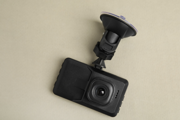 A black car dashcam with its suction cup mounting bracket attached, lying on a plain, light-colored textured surface.