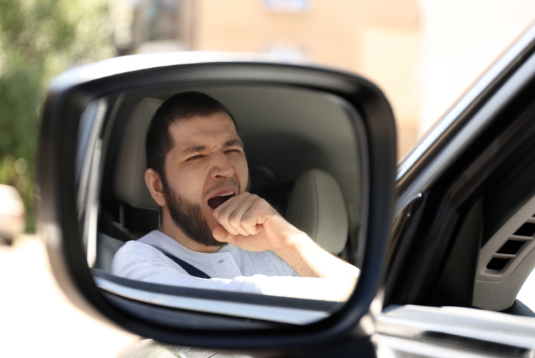 A reflection in a car's side-view mirror shows a man in the driver's seat yawning and covering his mouth with his hand.