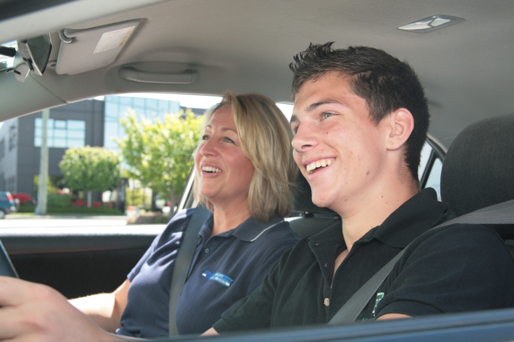 a young man is in the driver's seat with a blonde woman sitting beside him in the passenger seat. Both people are smiling.