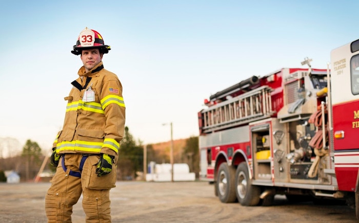 A firefighter in full tan turnout gear and a red helmet standing in front of a red fire truck during golden hour.