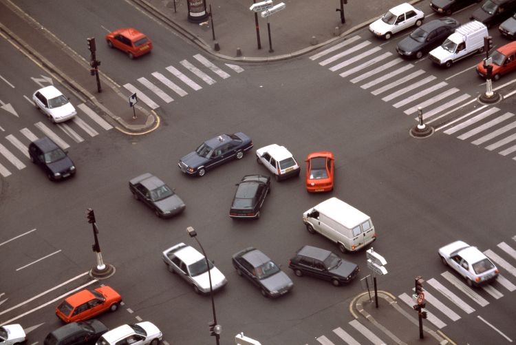 An aerial view of numerous cars and vans navigating a busy city intersection featuring zebra-striped crosswalks and traffic signals.