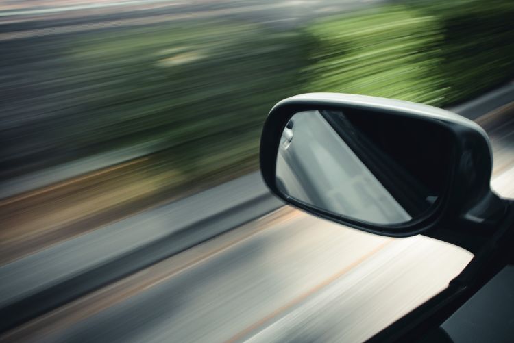 A close-up of a car's side-view mirror in sharp focus against a background of green trees and road, which are heavily motion-blurred to convey a sense of high speed.