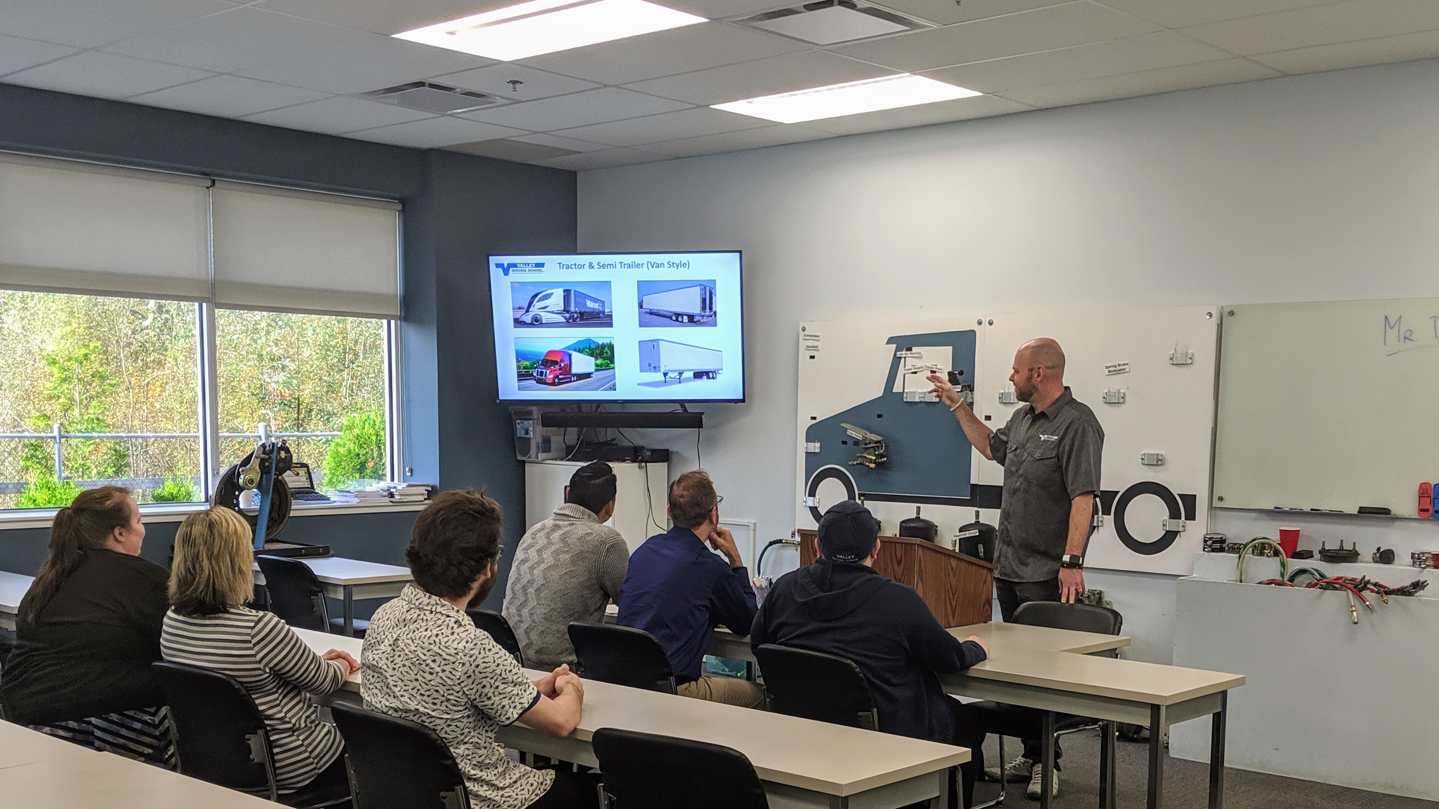 A Valley Driving School instructor teaching a commercial driving theory class using an interactive air brake board and a monitor displaying different truck types.