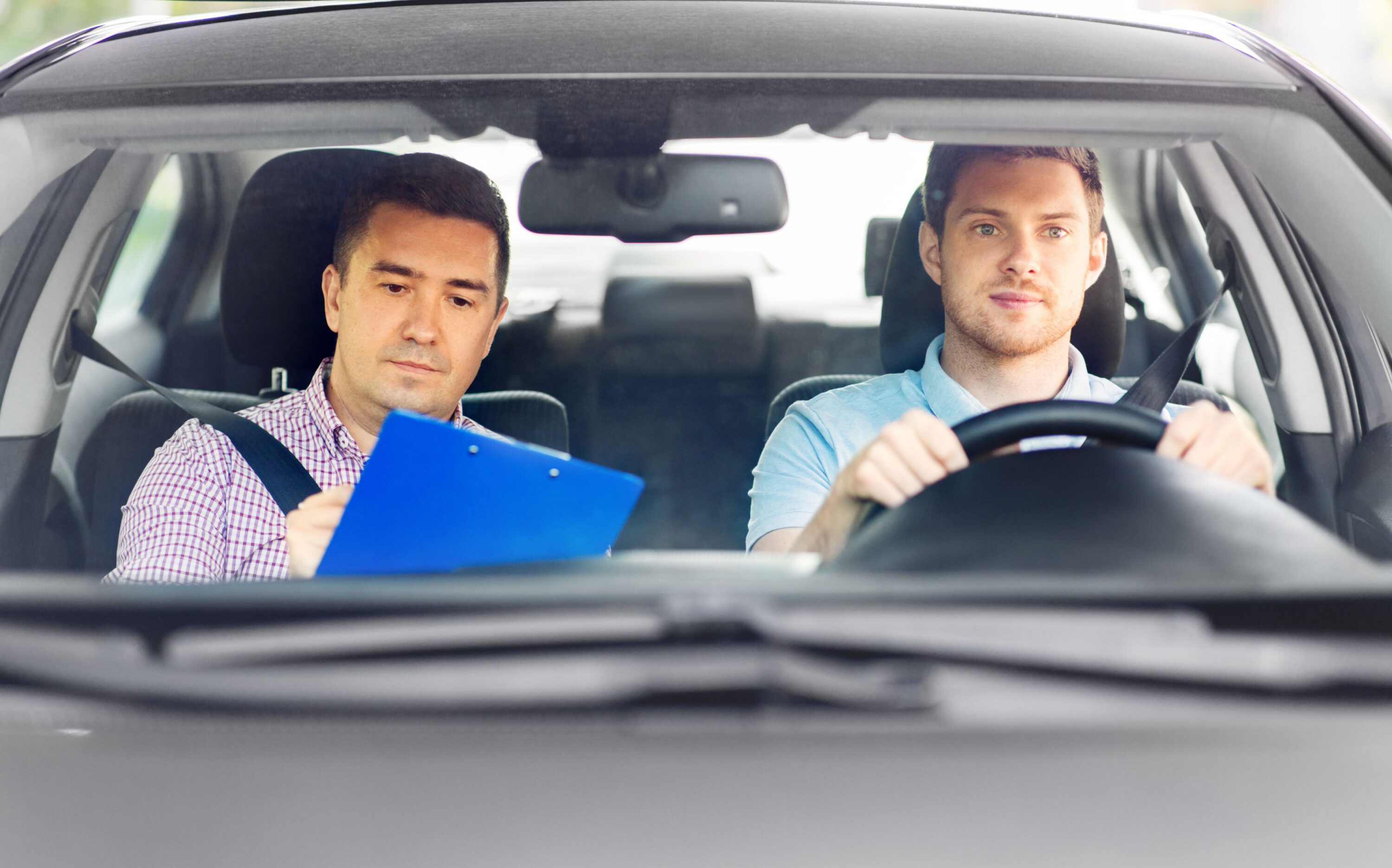 2 men behind in a car, one man is driving and another is marking a clipboard.