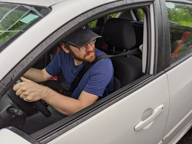 A male driver in a white car performing a shoulder check and looking over his shoulder while holding the steering wheel, demonstrating safe driving habits.