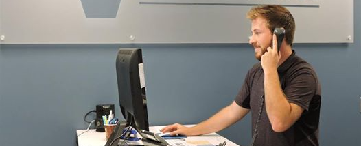 A young man stands at a computer desk in an office while answering a phone.