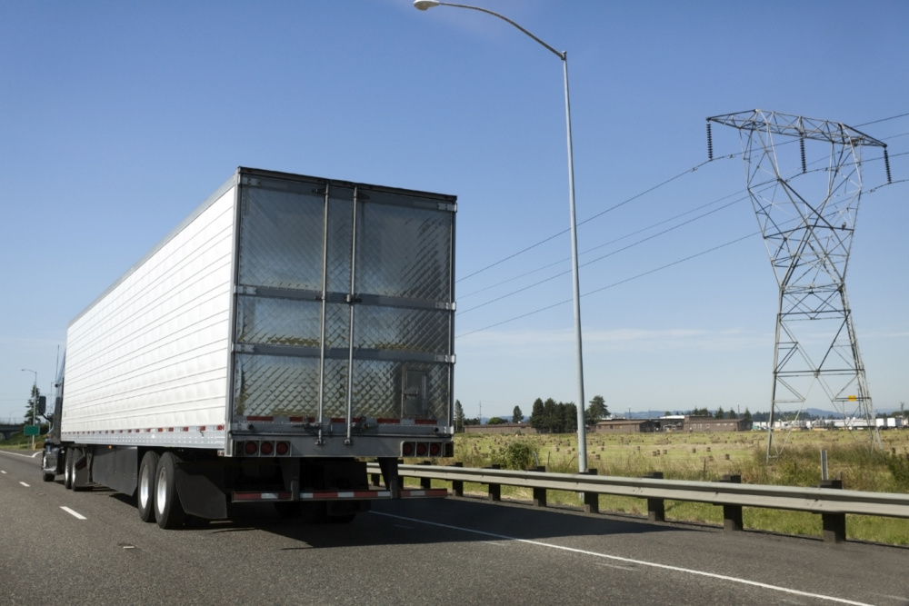 A semi Truck driving on the road beside farm land and power lines.