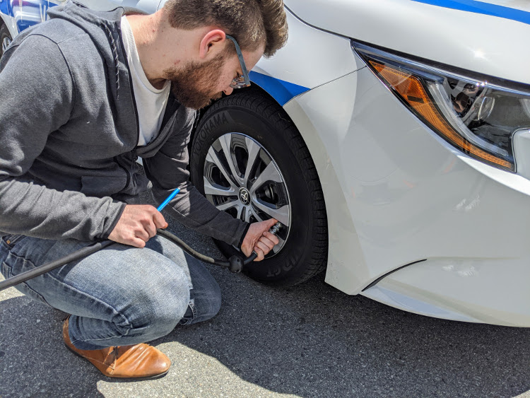 A Valley Driving School Instructor kneeling next to a white Toyota Corolla training vehicle to check and adjust the tire pressure using an air hose.