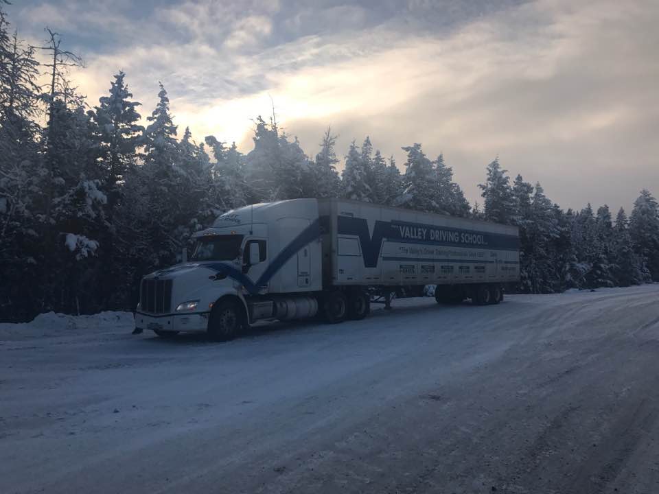A Valley Driving School truck and trailer parked on a snow-covered roadside with winter trees in the background.