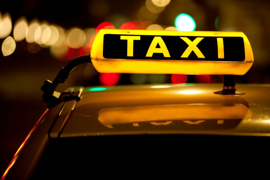 A glowing yellow "TAXI" sign on top of a car roof at night, reflecting on the vehicle's surface against a background of blurred, colorful city lights.