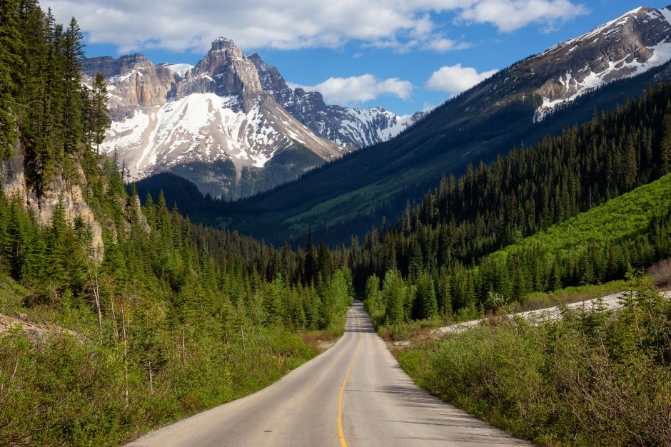 A paved two-lane highway stretches through a lush green valley toward towering, snow-capped mountain peaks under a bright blue sky with scattered white clouds.