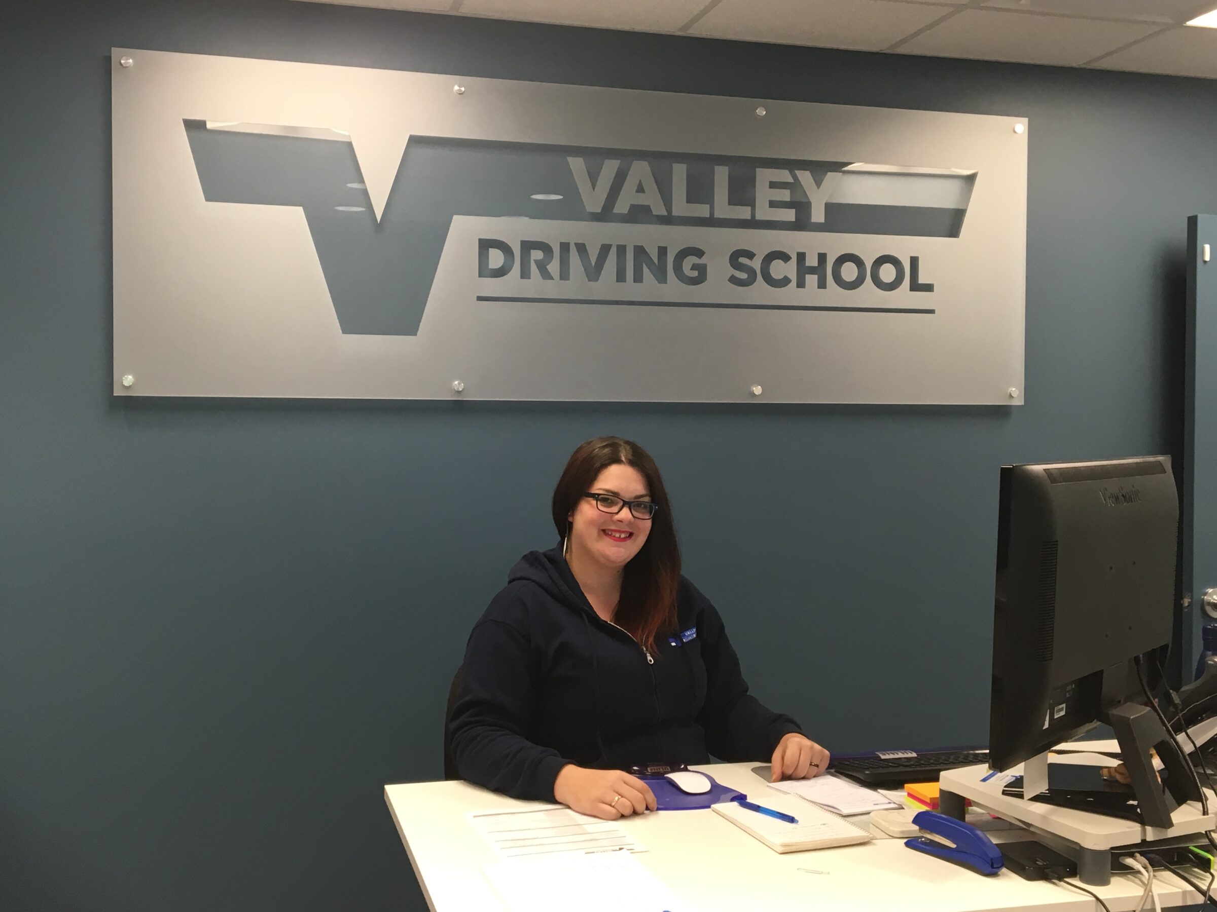 a young woman sits behind an office desk with a Valley Driving School sign on the blue wall behind her.