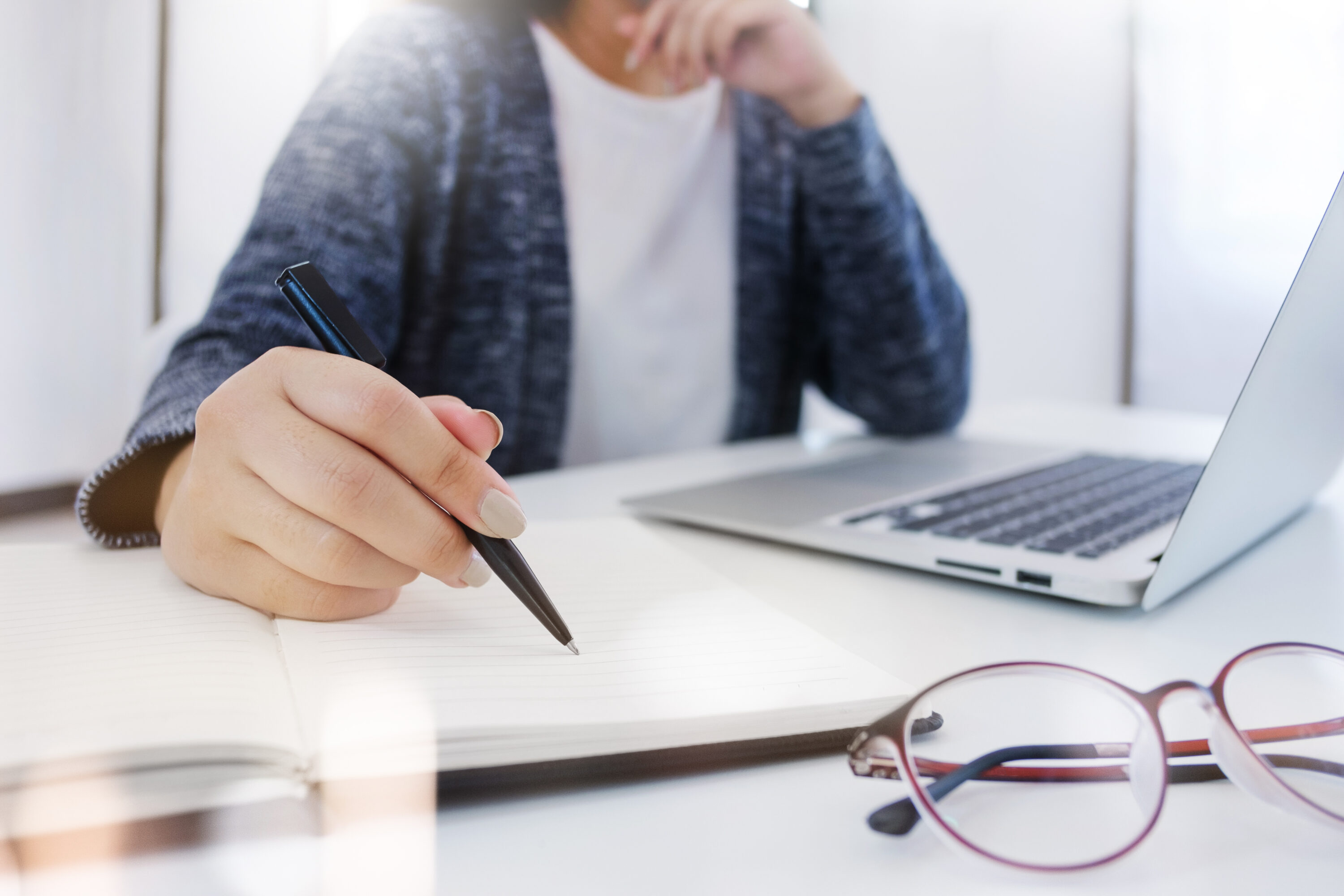 a person sitting at a white desk with a silver laptop. They are writing in a note book with a pair of glasses beside the notebook.