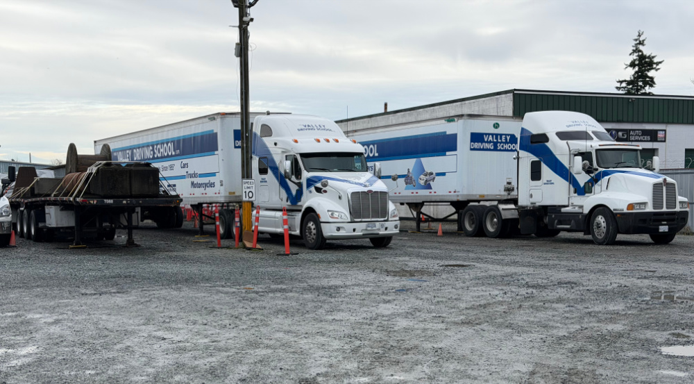 2 Valley Driving School tractor trailer units parked in a gravel yard, beside a flat deck trailer with various loads tied down.