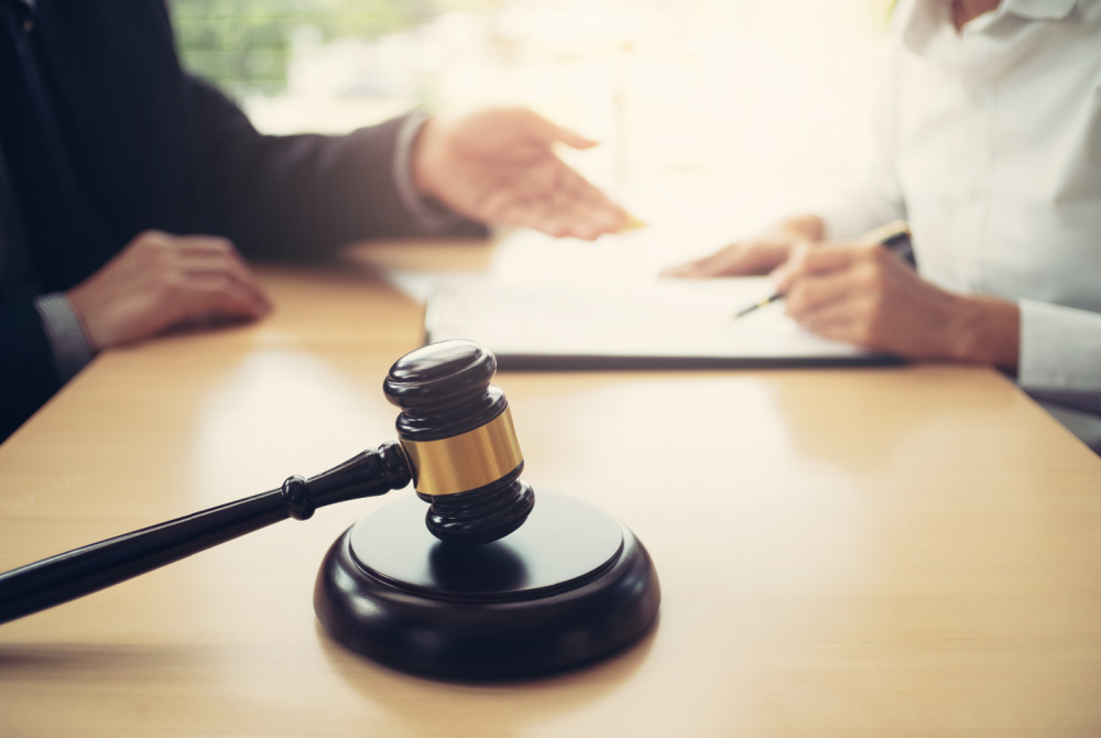 A lawyers office with 2 people at a desk. One person is writing a notebook and there is a gavel on the desk.