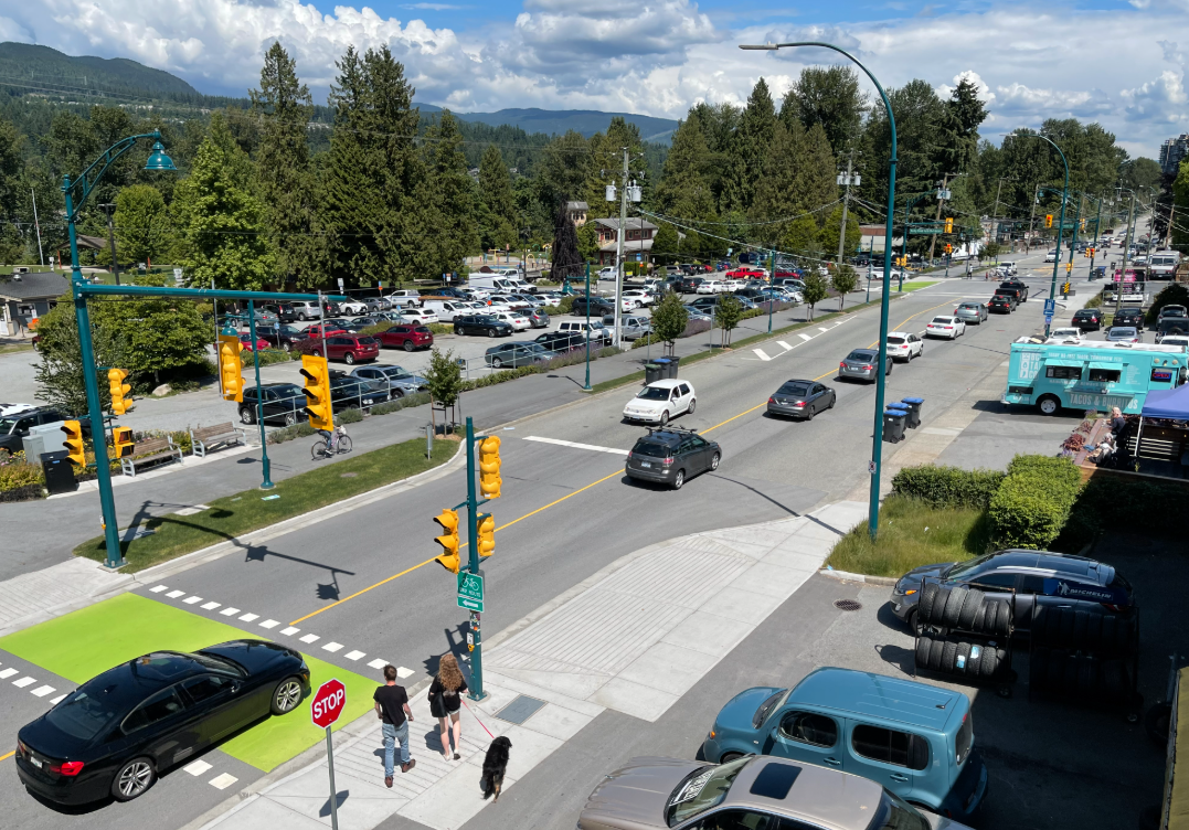 A busy road in Port Moody, BC with cars travelling on either side of the road. A black car is passing over a green crosswalk while 2 pedestrians with their dog wait to cross.