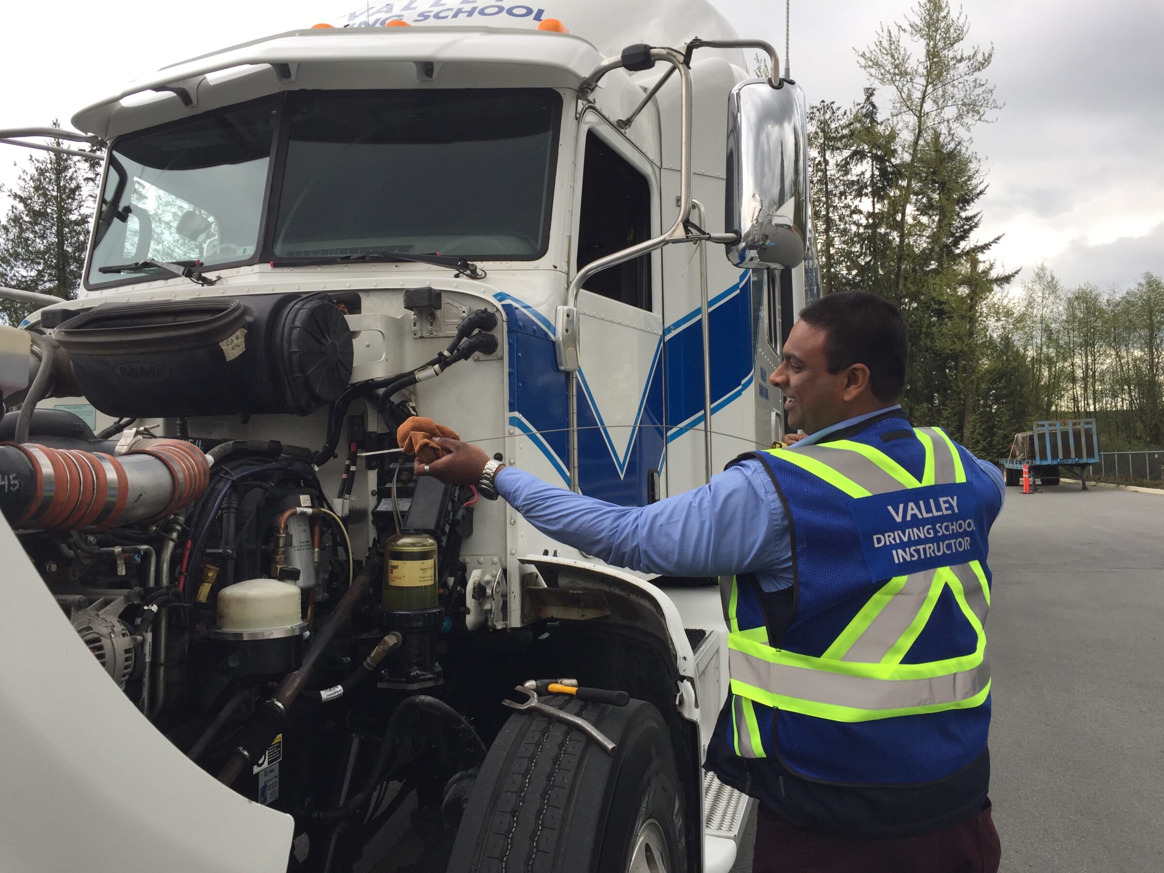 A Valley Driving School instructor in a blue safety vest performing a pre-trip inspection on a large white Valley Driving School truck.