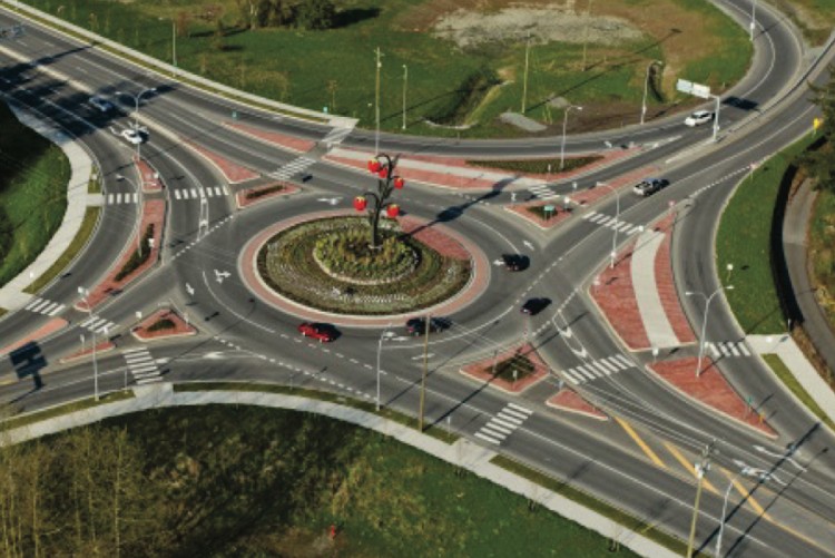 An aerial view of a multi-lane roundabout connecting several roads, surrounded by green trees and grassy areas.
