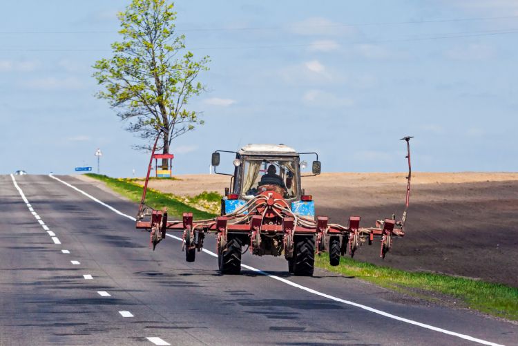 A blue tractor pulling agricultural equipment down a paved road, viewed from the perspective of a vehicle following closely behind.