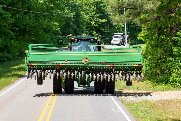 A green tractor pulling a large piece of farming equipment down a narrow, tree-lined rural road, with a view from the perspective of a vehicle following behind it.