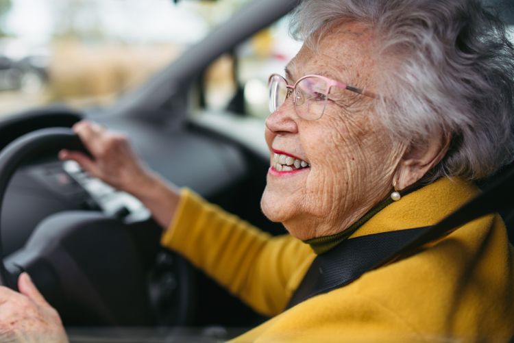 This image shows an older woman sitting in the driver's seat of a car, holding the steering wheel with a smile as she looks ahead out the front windshield.