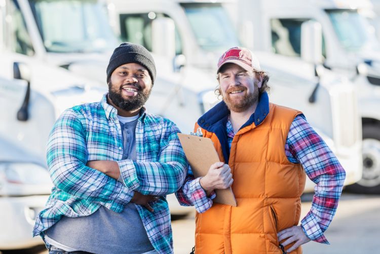 Two men wearing plaid shirts and hats standing side-by-side outdoors, smiling at the camera in front of a row of parked white semi-trucks, with one man holding a clipboard.