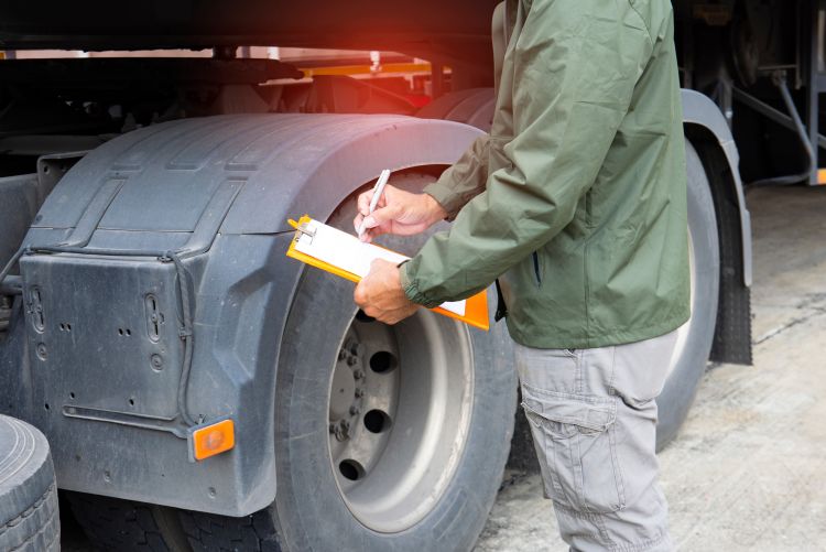 A close-up view of a person wearing a green jacket standing beside a large semi-truck tire, writing on a document attached to an orange clipboard.