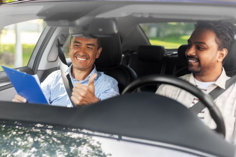 A photo of a driving examiner or instructor sitting in the passenger seat of a vehicle holding a clipboard, alongside a male driver behind the steering wheel.