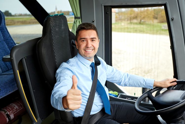 A photo of a smiling male driver wearing a light blue shirt and a tie, seated behind the steering wheel of a shuttle bus with his seatbelt fastened.