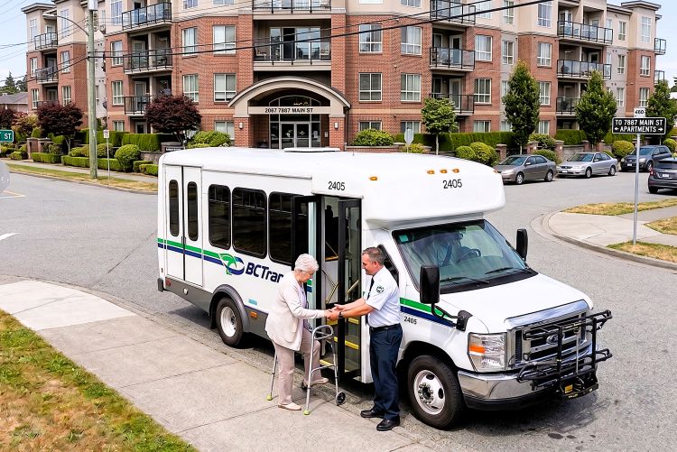 A photo of a white commercial shuttle bus parked on the street in front of a multi-story residential brick building.