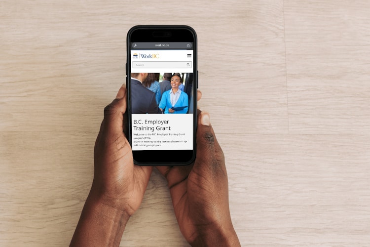A person holding a smartphone that displays the WorkBC BC Employer Training Grant webpage.