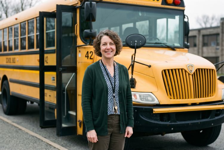 A smiling woman with long brown hair standing outside next to an open door of a yellow school bus.