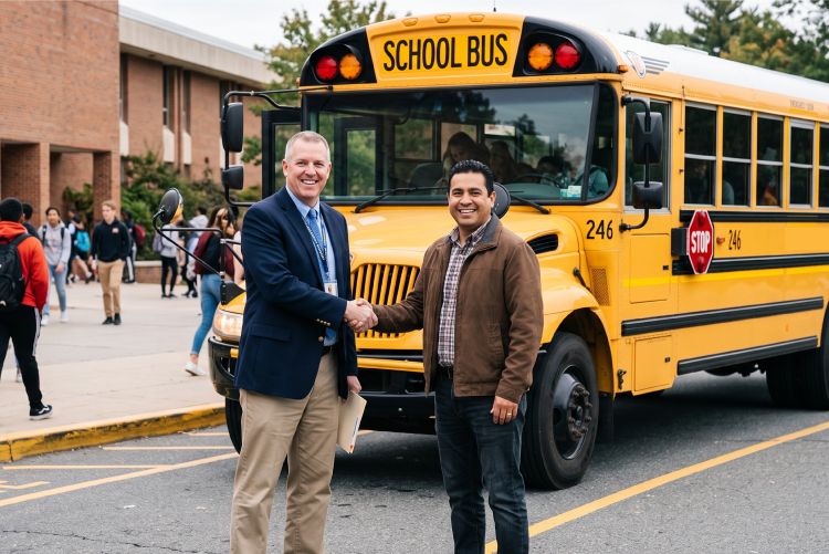 A school bus driver shaking hands with the school principle. There is a yellow school bus and a school building in the background.