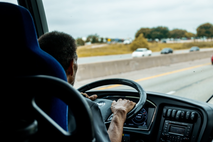 A view looking past the driver's side mirror of a school bus, showing a male bus driver sitting in the driver's seat.