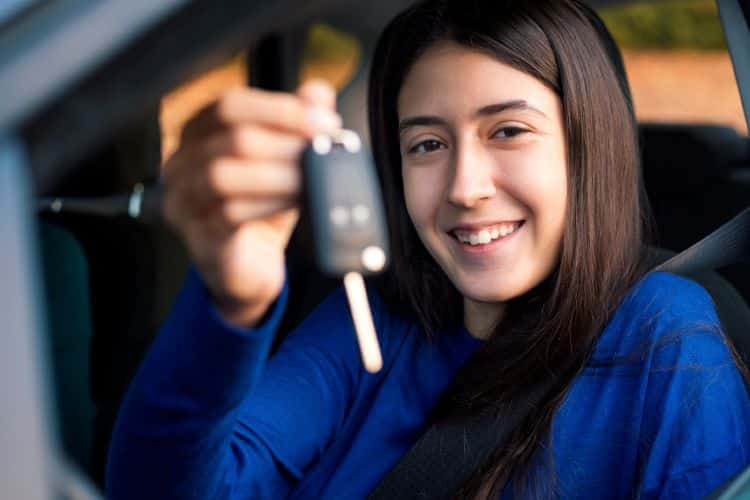 A smiling woman sitting inside a vehicle holding up a set of car keys while sitting in the driver's seat.