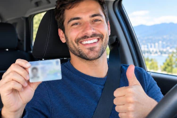 A man sitting in the driver's seat of a car, smiling while holding up his driver's licence.