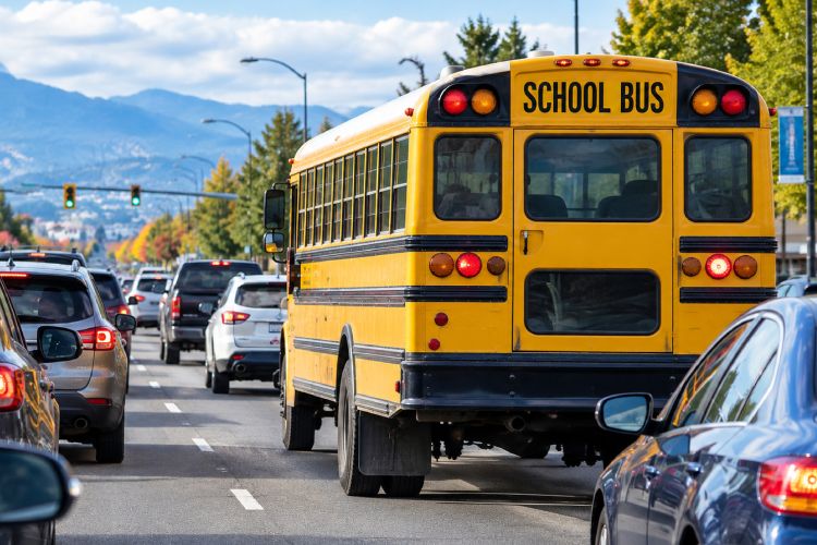 A yellow school bus driving down a multi-lane city road with traffic, buildings, and streetlights in the background.
