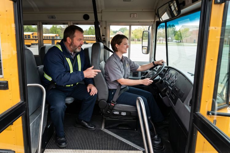 A woman learning to drive a school bus, sitting at the driver's seat, with an Instructor sitting behind her, coaching.